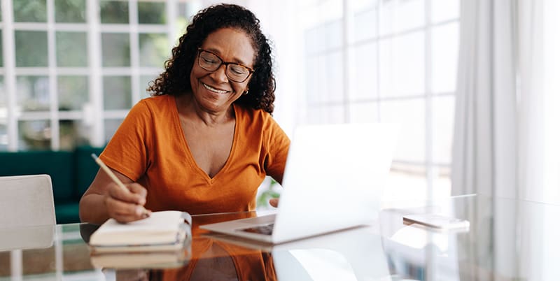 Happy senior woman sitting at a table in her home office, drafting her last will and testament in a journal and taking care to allocate her assets. Retired woman leaving a plan in place for the future.